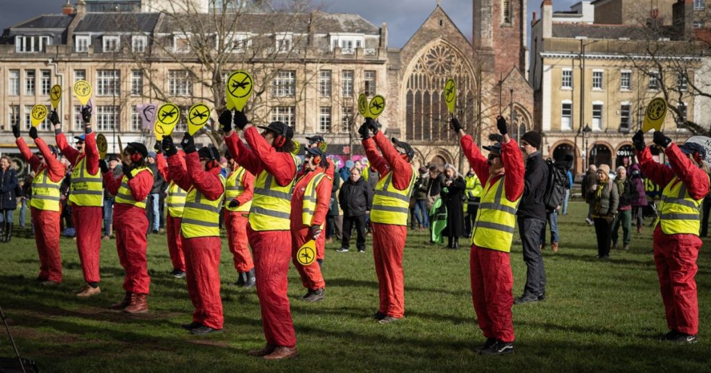 A group of 12 protesters dressed as air traffic controllers in high-vis vests holding up paddles with a vector of an airplane crossed out, stand on College Green, campaigning against the expansion of Bristol Airport. Several members of the public stand behind them looking on to the protest.